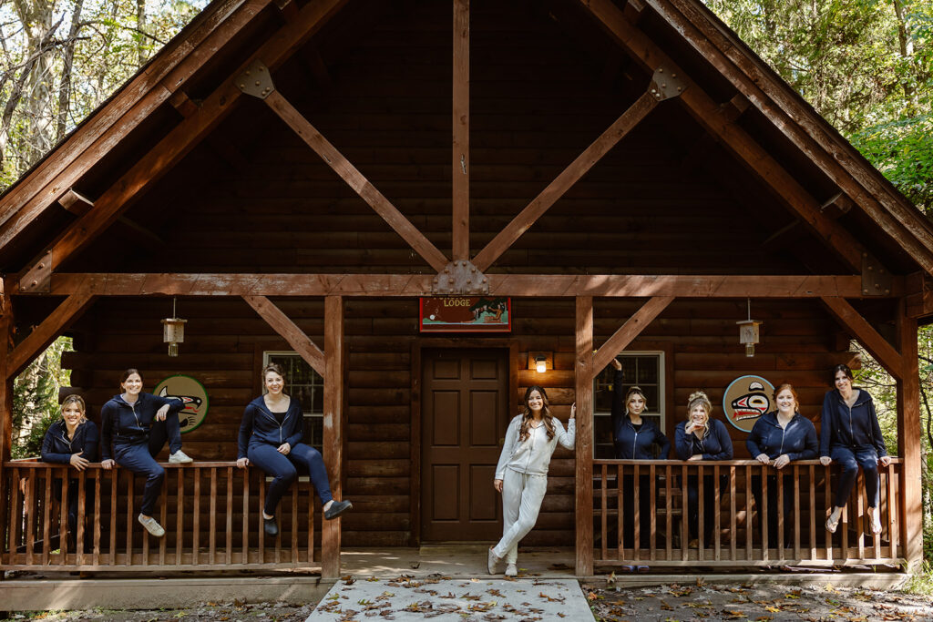 A bride and her bridesmaids all in matching sweatsuits are posing in front of A-frame cabin, The bride is wearing a white sweatsuit, and the other woman are wearing blue.