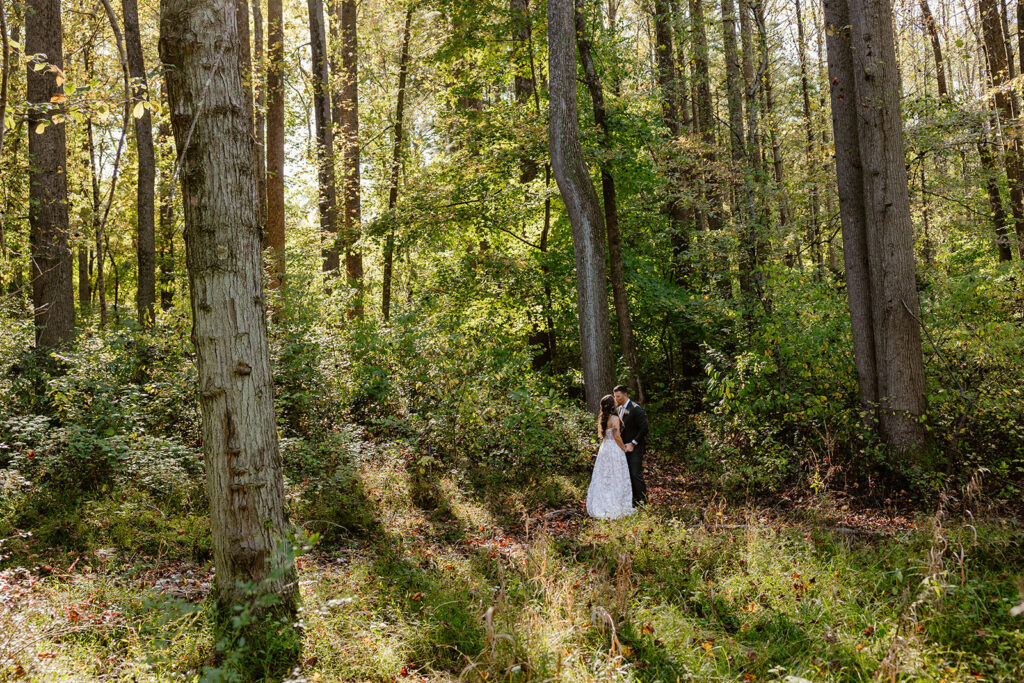 A bride and groom stand in the forest while holding hands on their wedding day. They are facing each other and kissing