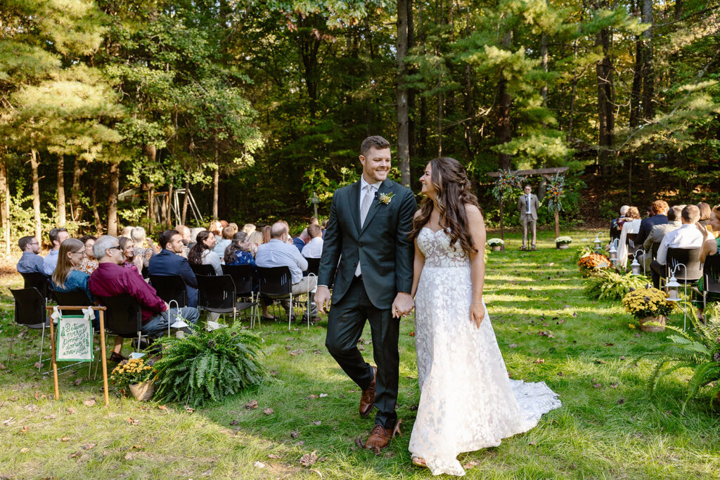 A bride and groom walking down the aisle during the recessional of their ceremony. They are holding hands and smiling at each other. They are in the woods with their closest family and friends. camp puh'tok wedding