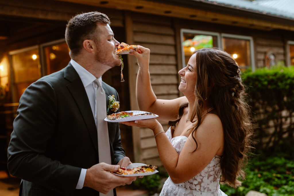 A bride feeds her groom a slice of pizza like a couple would feed each other cake on their  wedding day. The bride is laughing while she feeds him