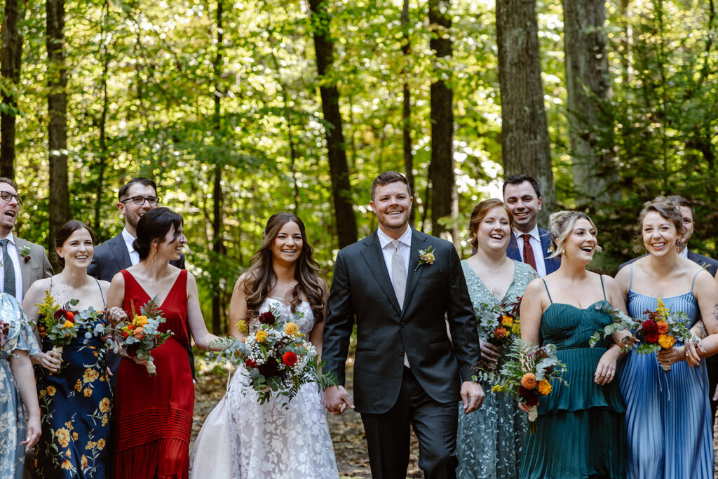 A couple walks in the forest with their closest friends on their wedding day. Everyone is laughing and smiling, and the bride and groom are holding hands