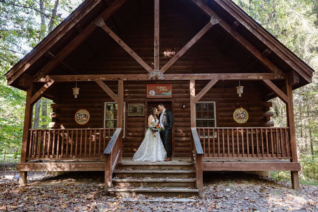 A couple stands in front of a cabin at a summer camp on their wedding day. They are facing one another and smiling. The bride is wearing a lace dress, and the groom is wearing a suit. camp puh'tok wedding