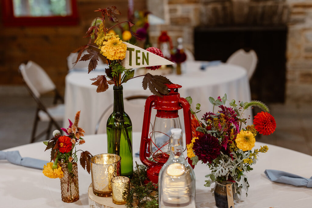 A photo of the details that make up the table scape at a summer camp wedding. The tables have red lanterns, wild flowers, and pennants with the table numbers.