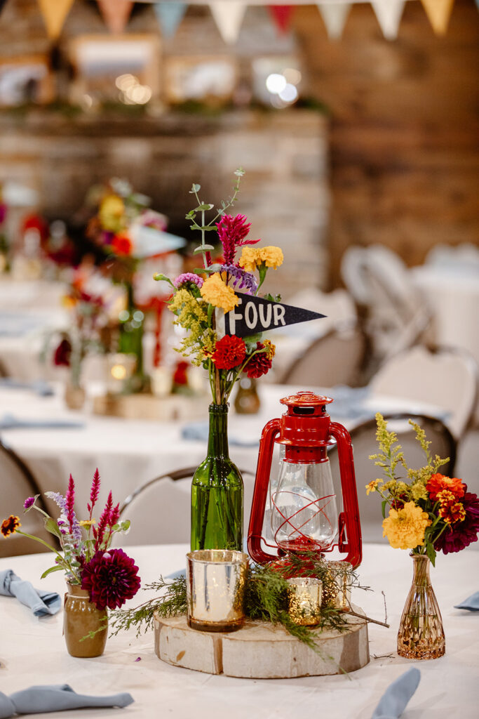 A photo of the details that make up the table scape at a summer camp wedding. The tables have red lanterns, wild flowers, and pennants with the table numbers. camp puh'tok wedding