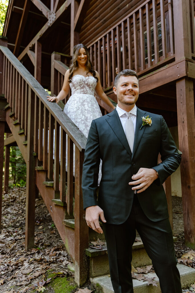 A groom is standing at the bottom of the steps of an a-frame cabin while his bride to be walks down them during their first look. You can see the excitement in both of their faces.