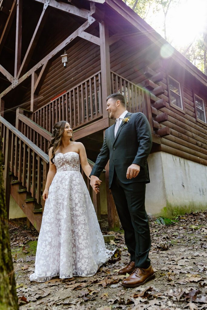 A bride and groom stand in front of am A-frame Cabin at a campground holding hands after seeing each other for the first time. The bride is wearing a lace dress, and the groom is wearing a suit