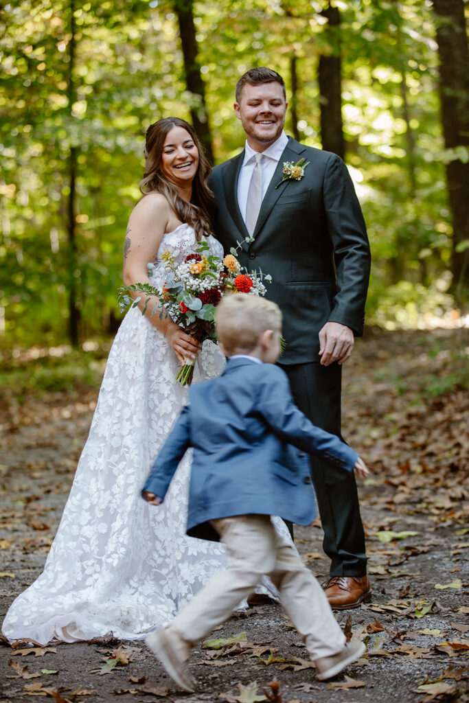 A bride and groom stand in the forest. The bride is wearing a lace strapless bride and holding a bouquet. There is a ring bearer running around the couple, and they are smiling