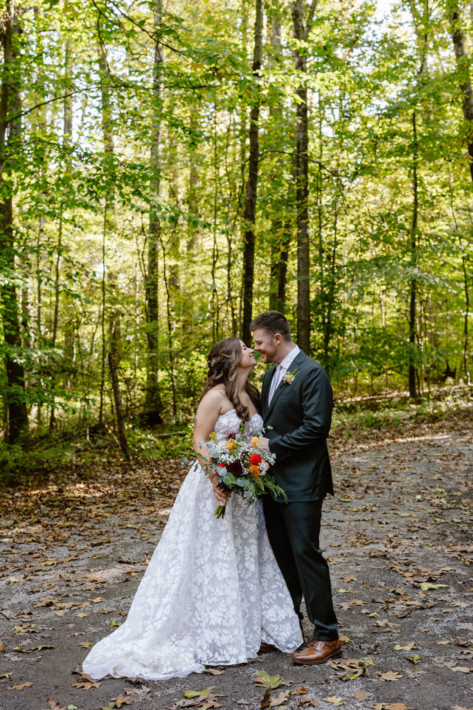 A bride and groom stand in the forest looking at each other and smiling, The bride is wearing a lace strapless bride and holding a bouquet