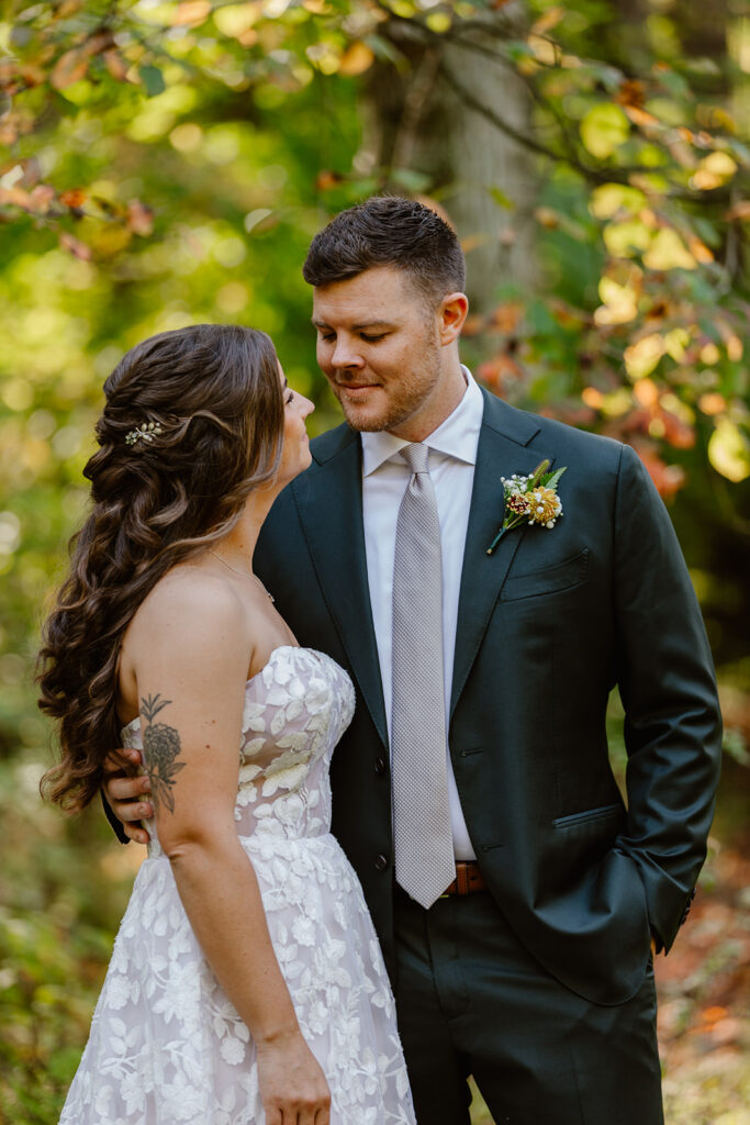 A groom looks lovingly at his bride on their wedding day. They are standing in the forest. She is wearing a strapless dress