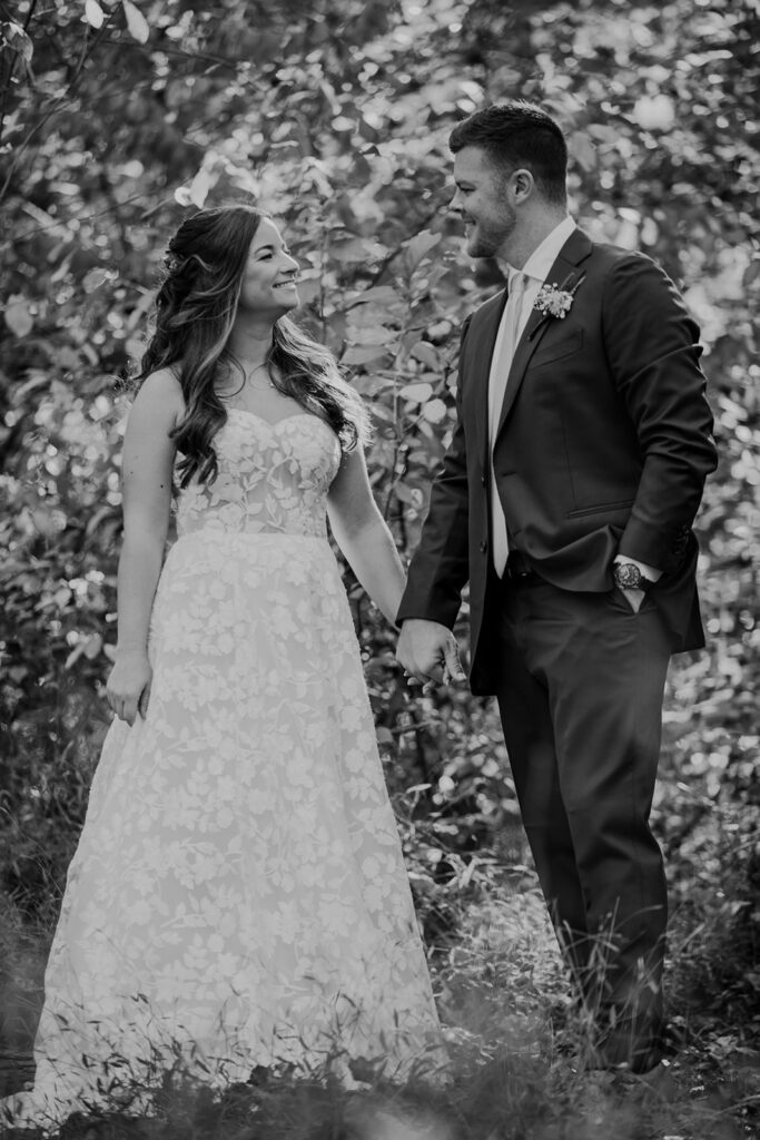 A black and white photo  of a couple holding hands smiling at one another in the forest. She is wearing a strapless lace wedding dress, and he is wearing a suit