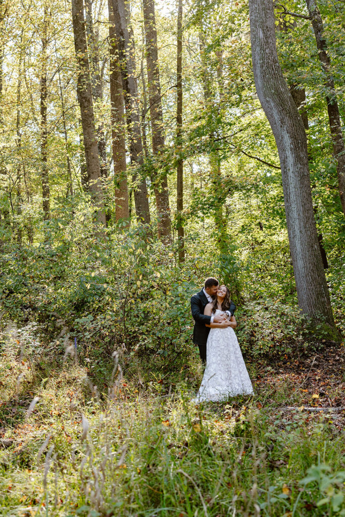 A bride and groom stand in the forest while holding hands on their wedding day. The groom is standing behind her, and kissing her cheek