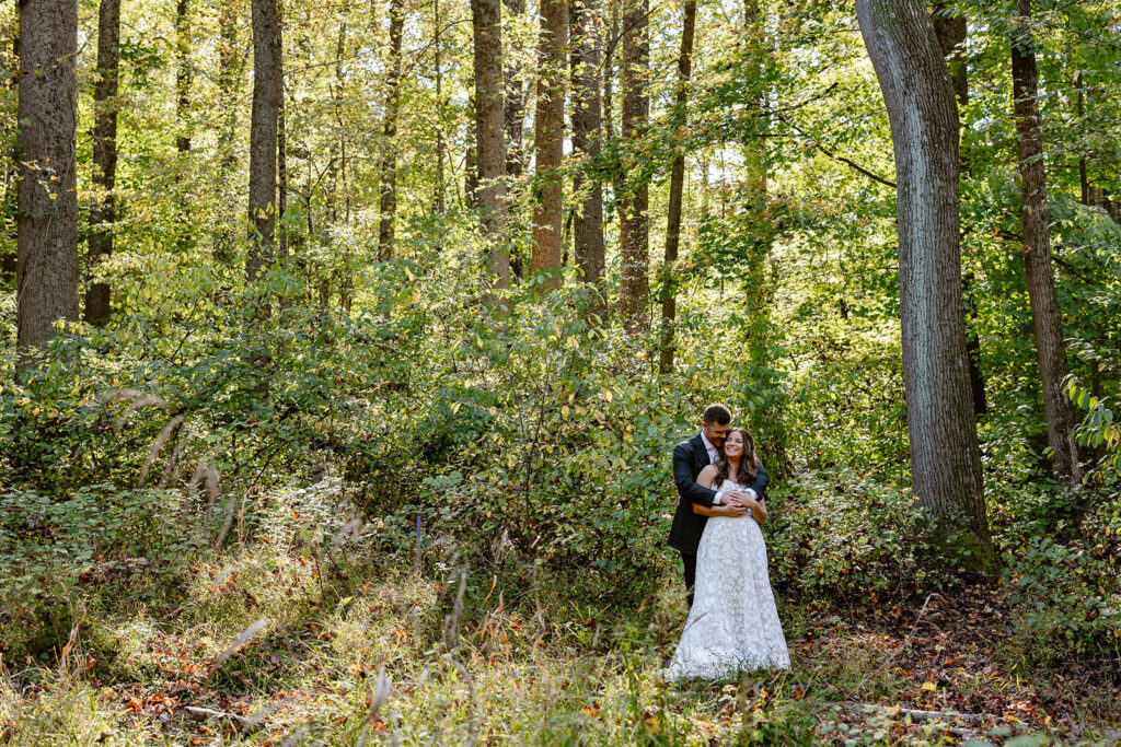 A bride and groom embrace in the woods. The groom is embracing the bride from behind her, and they are kissing.