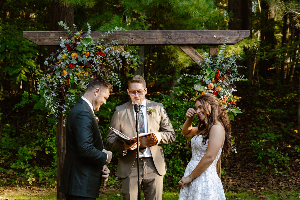 A bride and groom stand in front of a wooden alter adorned with flowers durning their wedding ceremony. The officiant is speaking and the bride is laughing while wiping away a tear