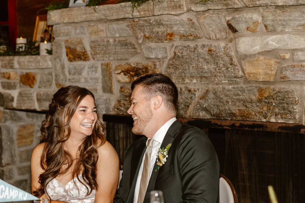 A bride and groom laughing at one another while listening to a toast from the father of the bride at their summer camp wedding.