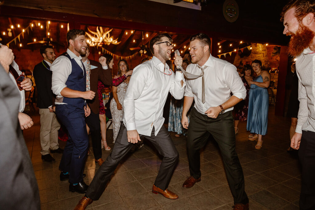 A groom and his friends being funny on the dance floor. The grooms friend is holding the grooms tie, and singing into it like it is a microphone.