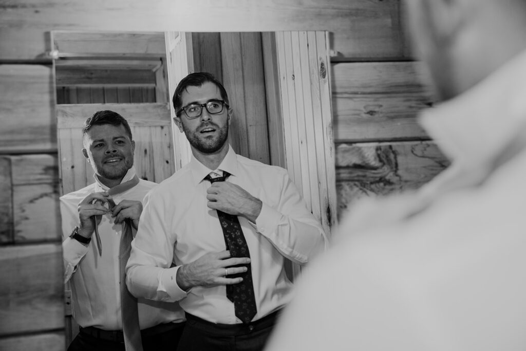 A groom and the officiant getting ready in a shared mirror. The men are both fixing their ties