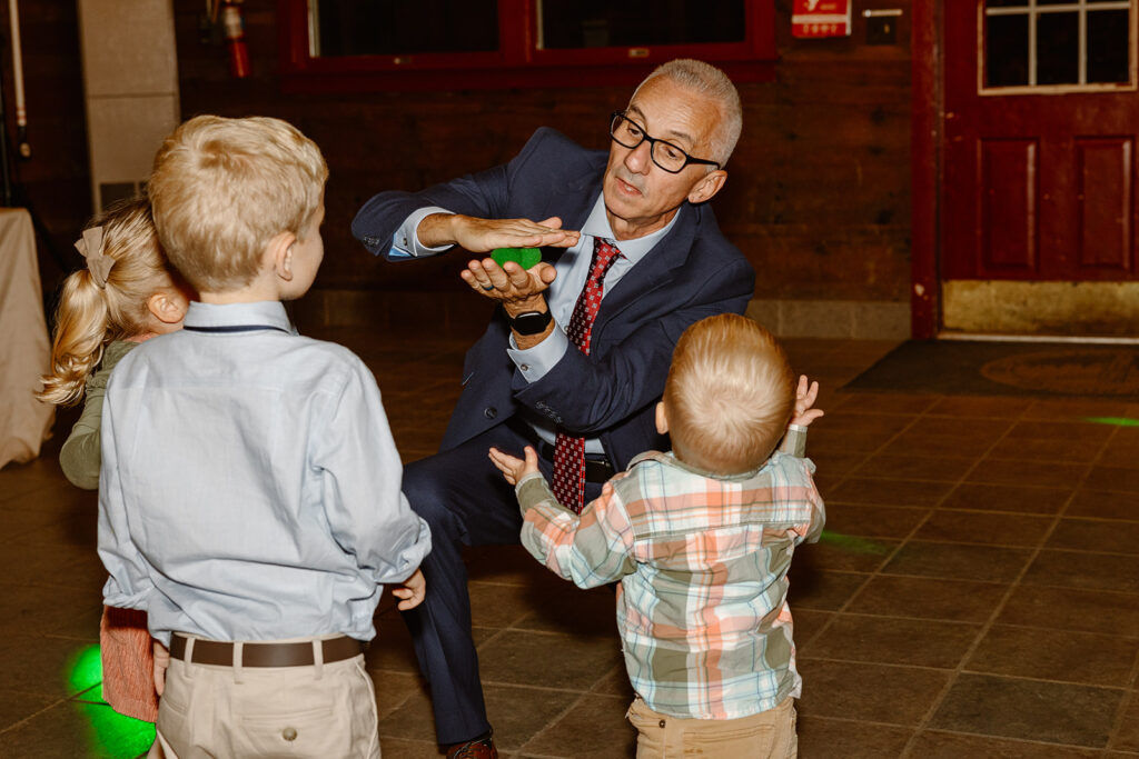 The bride's father performs magic for the kids at the wedding. He has a green foam ball that he makes disappear, and then reappear in the hand of a kid