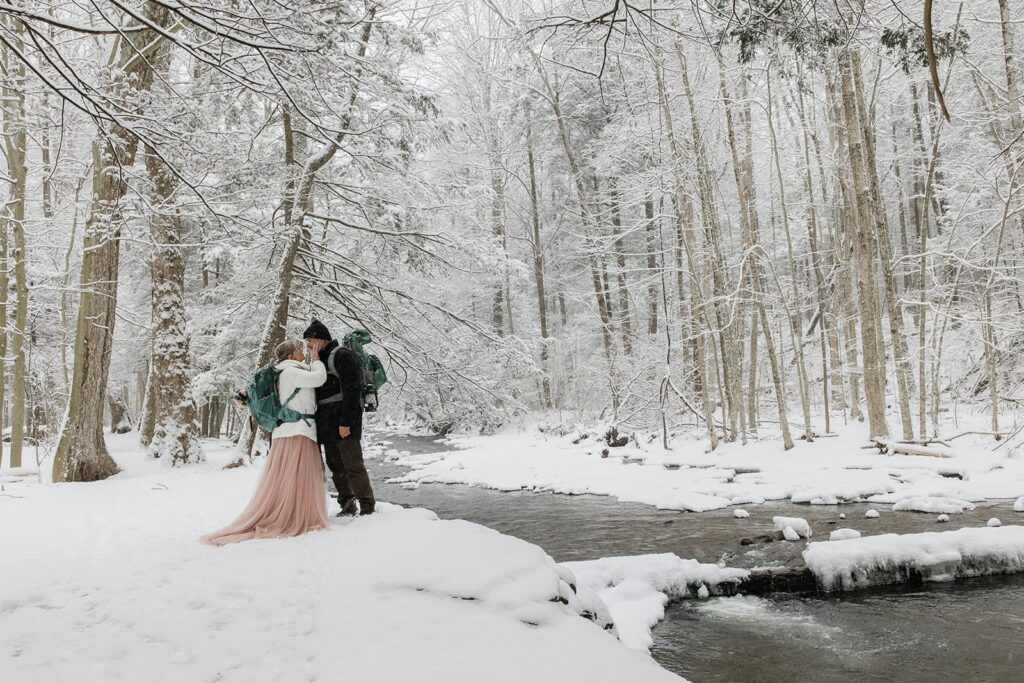 A couple stand in front of kitchen creek during their snowy elopement at one of the best places to elope in Pennsylvania, Ricketts Glen State Park