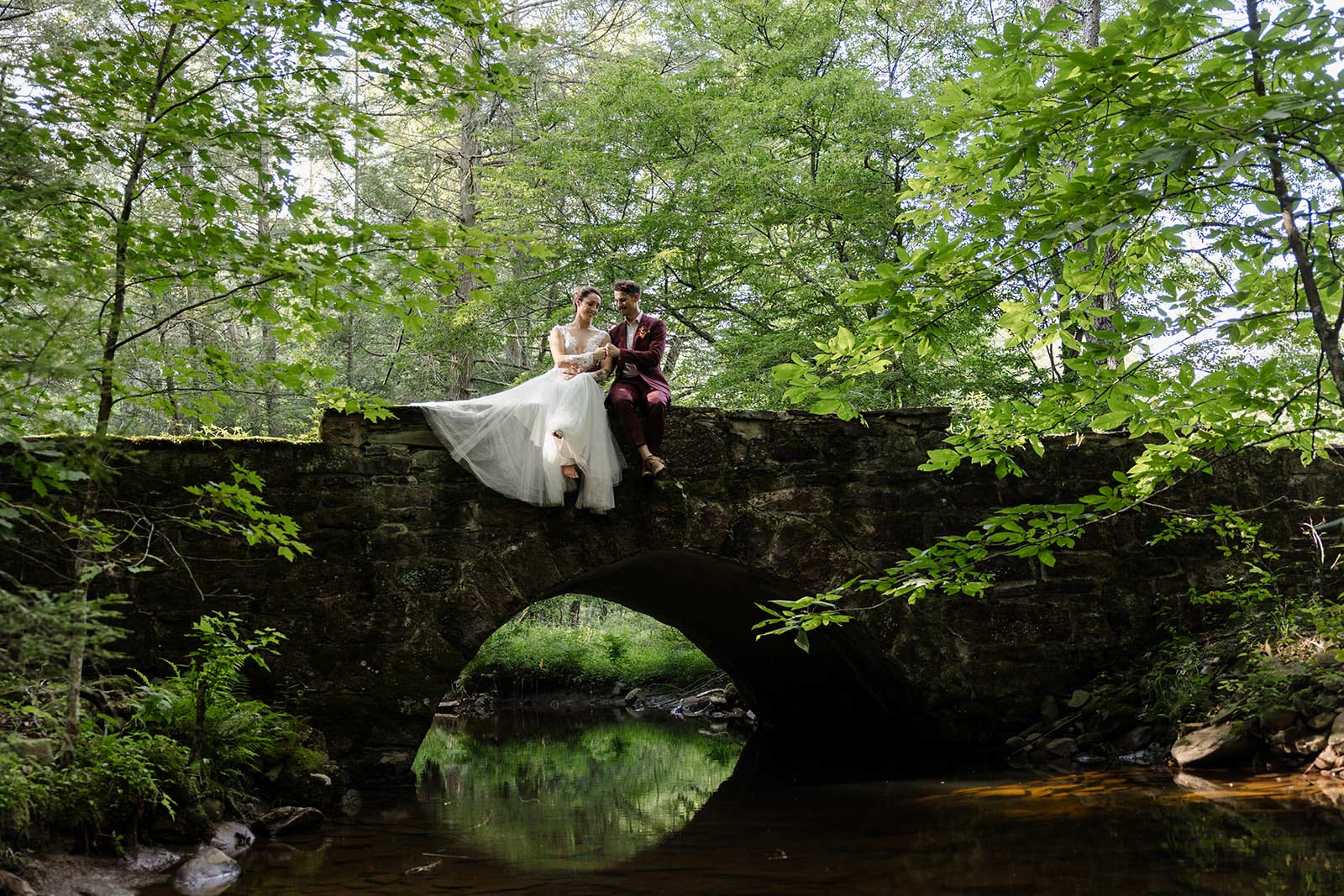 A bride and groom fist bump while sitting on a bridge near Ricketts Glenn, one of the best places to elope in Pennsylvania