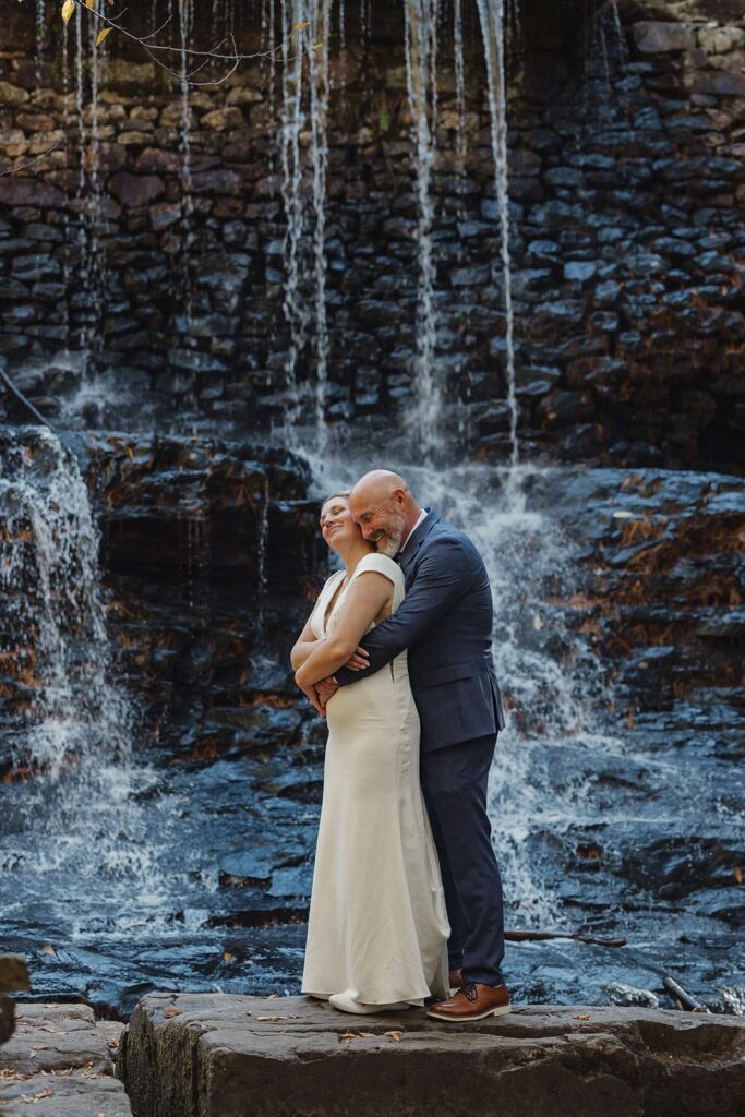 A groom snuggles behind his bride in front of Hawk Falls during their elopement 