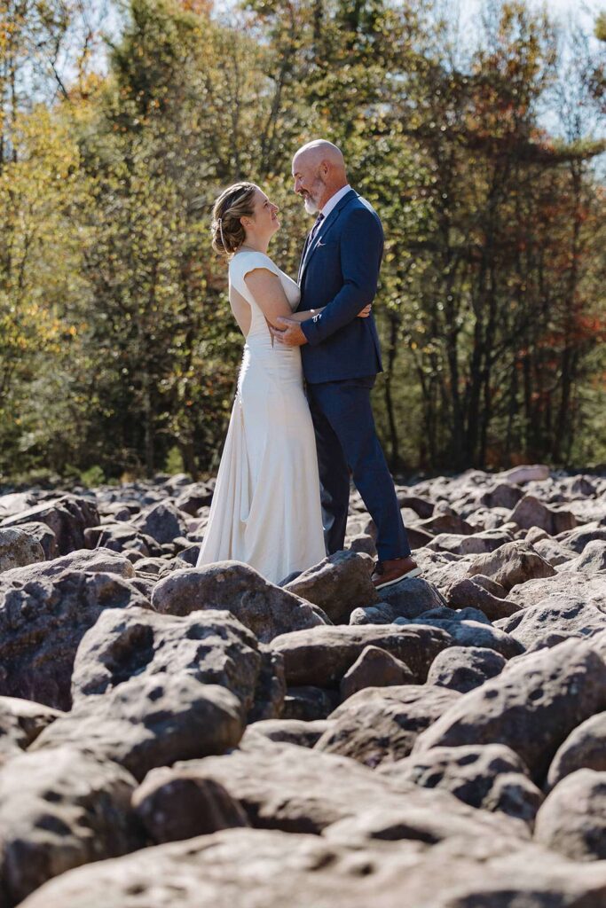 A bride and groom stand in the middle of a boulder field in Hickory Run State Park in Pennsylvania. They are looking at one another and smiling. 