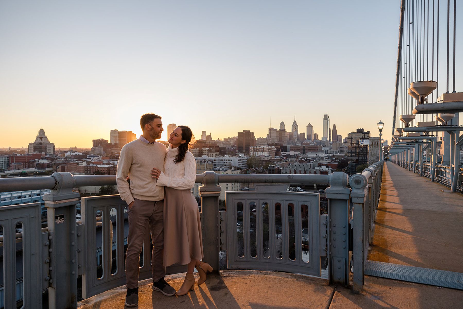 a couple standing next to each other on bridge at sunset the woman has her hand on the man's stomach and is looking up at him