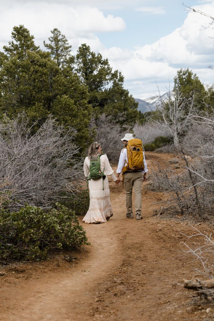 A bride and groom walking while holding hands. They are both wearing backpacks