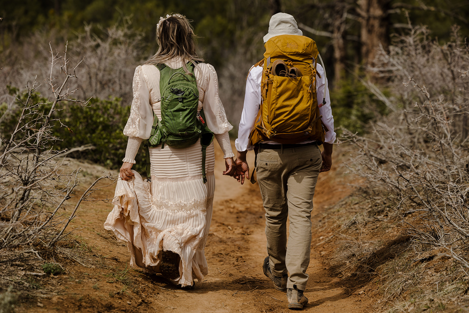 A couple walks hand in hand on a hike on their elopement day. They are both wearing their backpacks and hiking boots