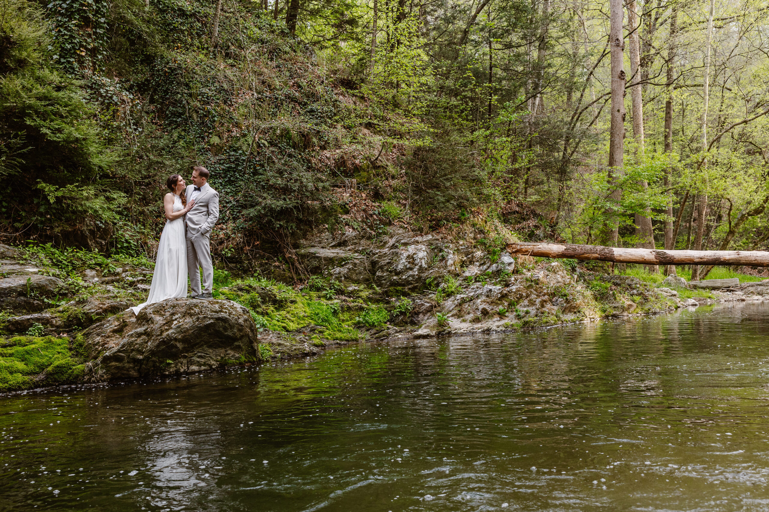 a couple standing on a rock together next to a river the woman has her hand on the man's chest the woman is also wearing a wedding dress the man is wearing a suit