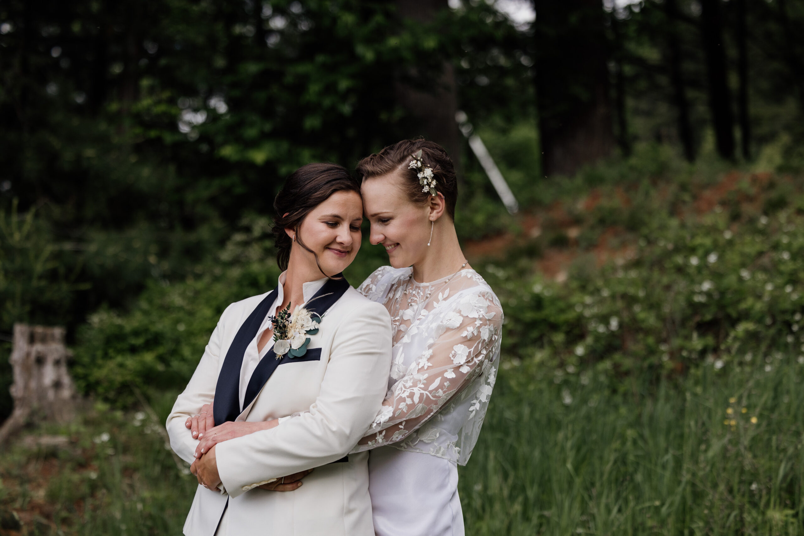 two women in the woods one woman is hugging the other woman from behind one woman is wearing a white wedding dress the other woman is wearing a white suit