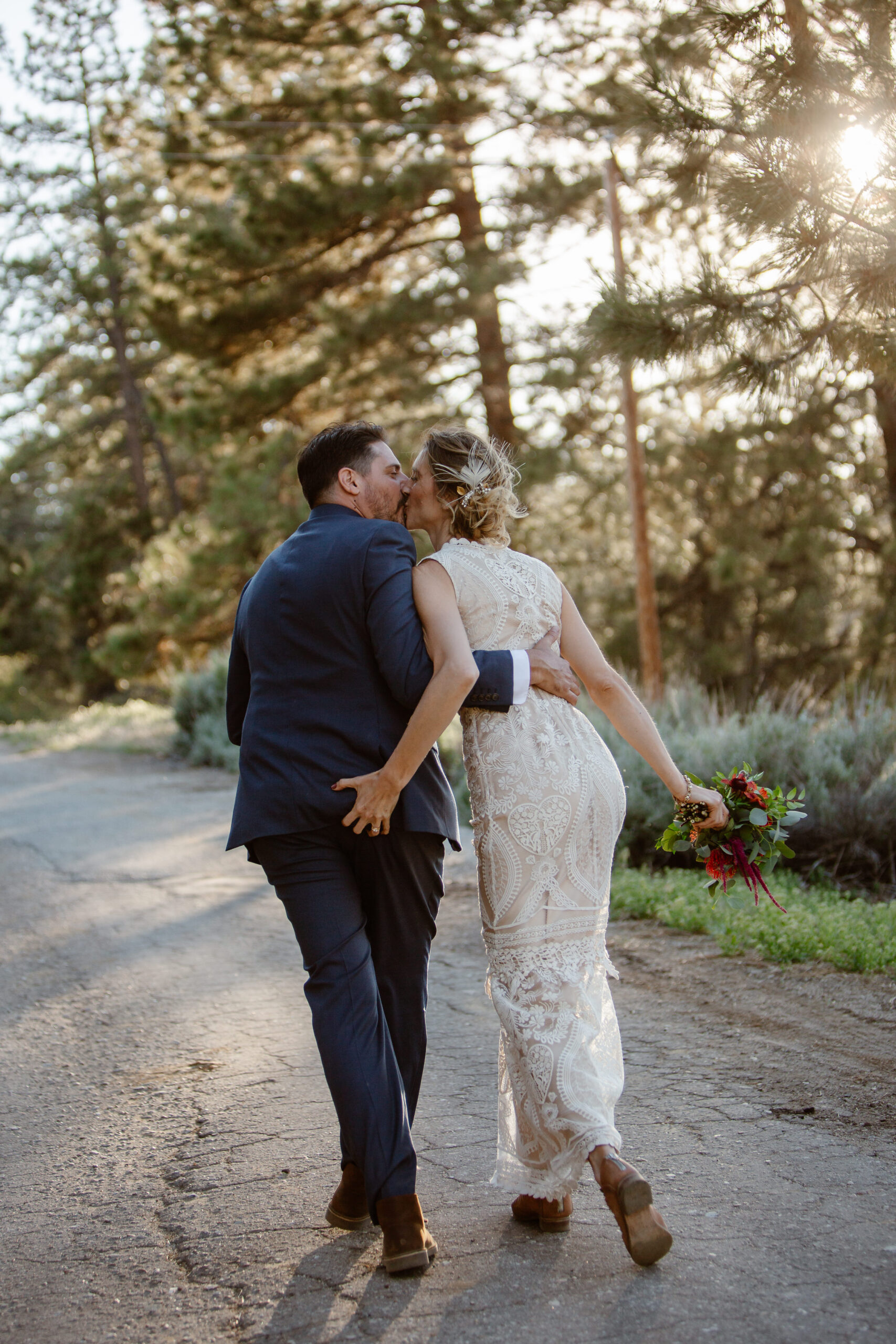 a couple kissing each other and walking down a path next to the woods the woman is waering a white dress the man is wearing a blue suit