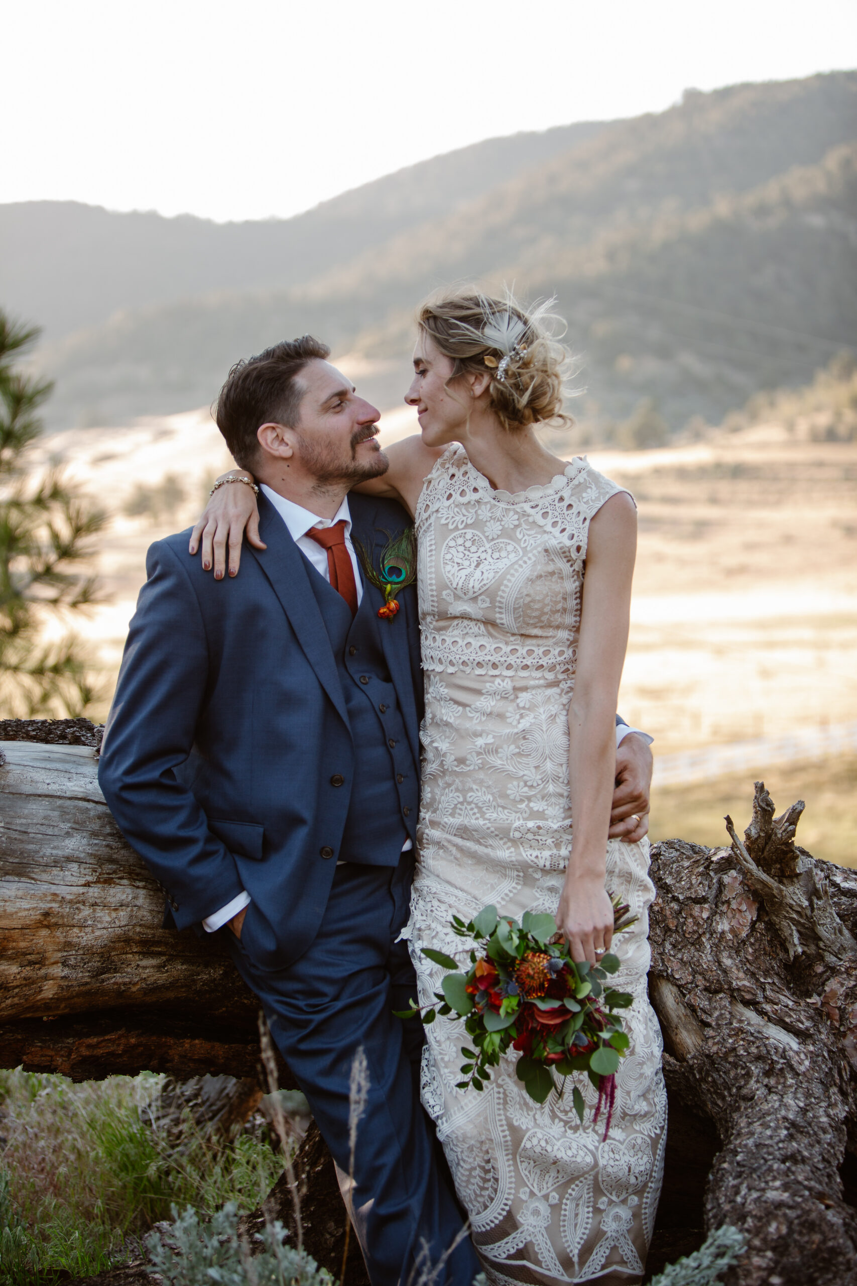 a couple sitting against a fallen tree looking at each other and smiling the woman is wearing a wedding dress and holding flowers the man is wearing a blue suit