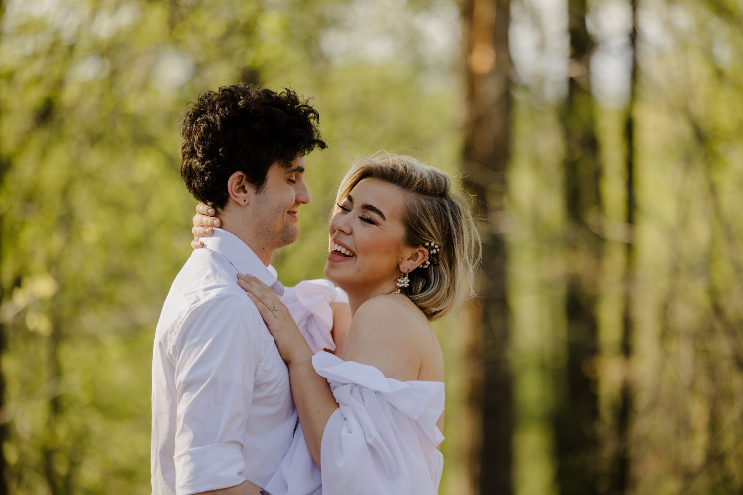 a couple embracing each other and smiling the woman is wearing a white dress the man is wearing a white button up they are in the woods