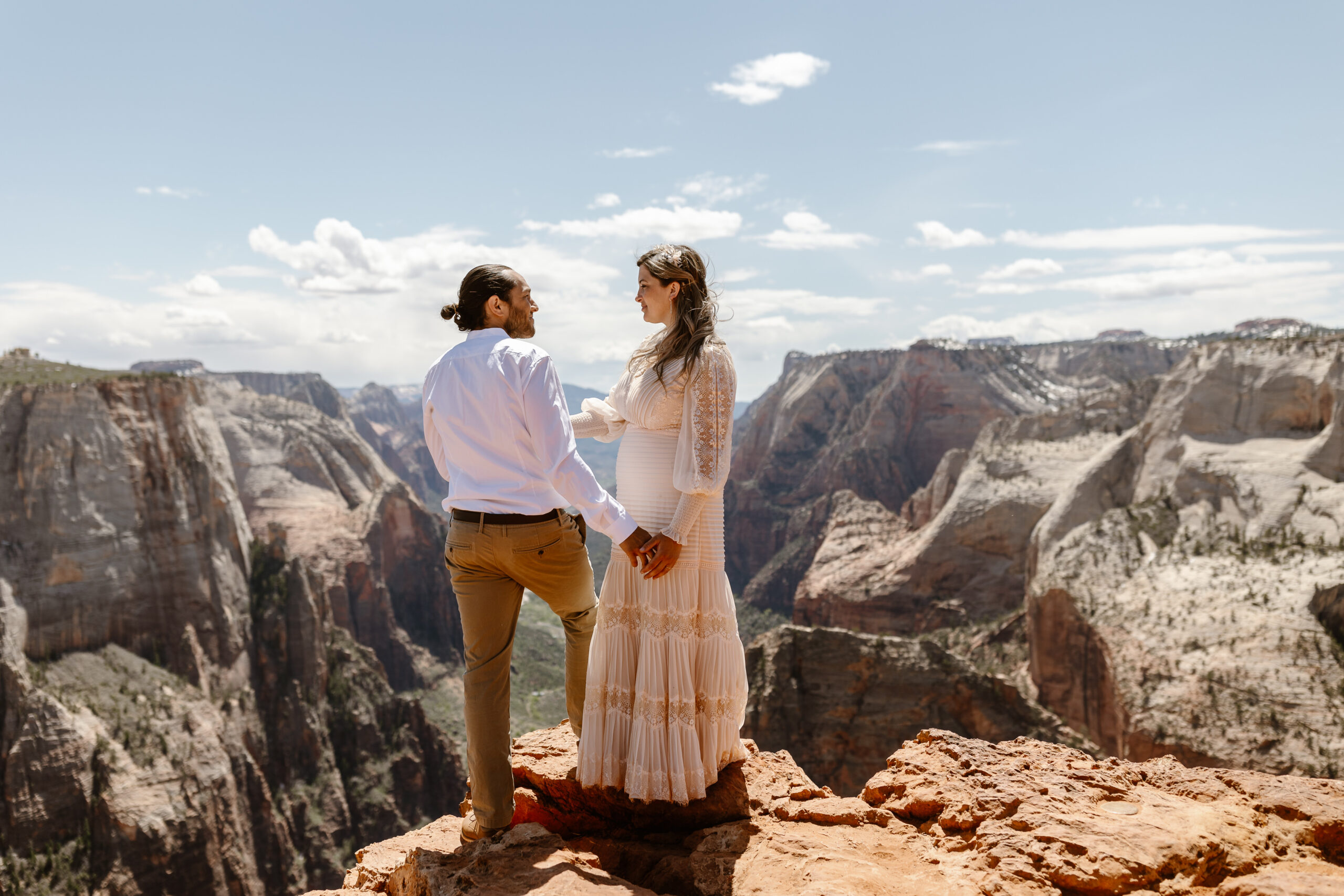 a couple standing together holding hands next to a valley the woman is wearing a white wedding dress the man is wearing a white button up and suit