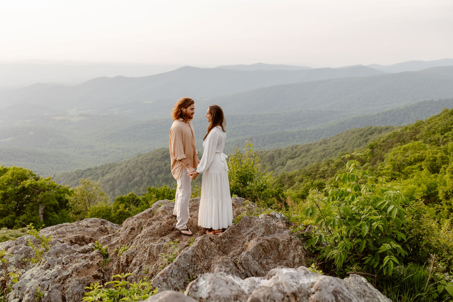 a couple standing on a rock together holding hands and smiling at each other behind them is a valley
