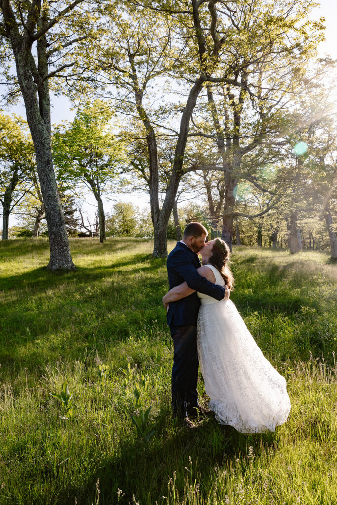 A bride and groom kiss in a grassy field while the sun backlights them