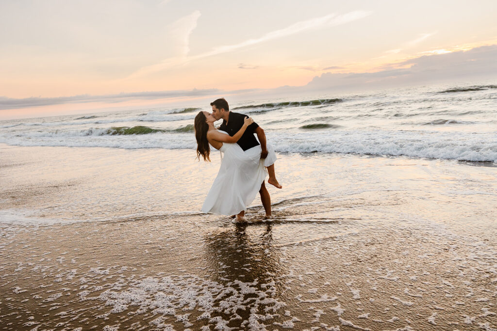 A groom dips his bride as the tide washes over them at Virginia Beach during their Virginia Elopement