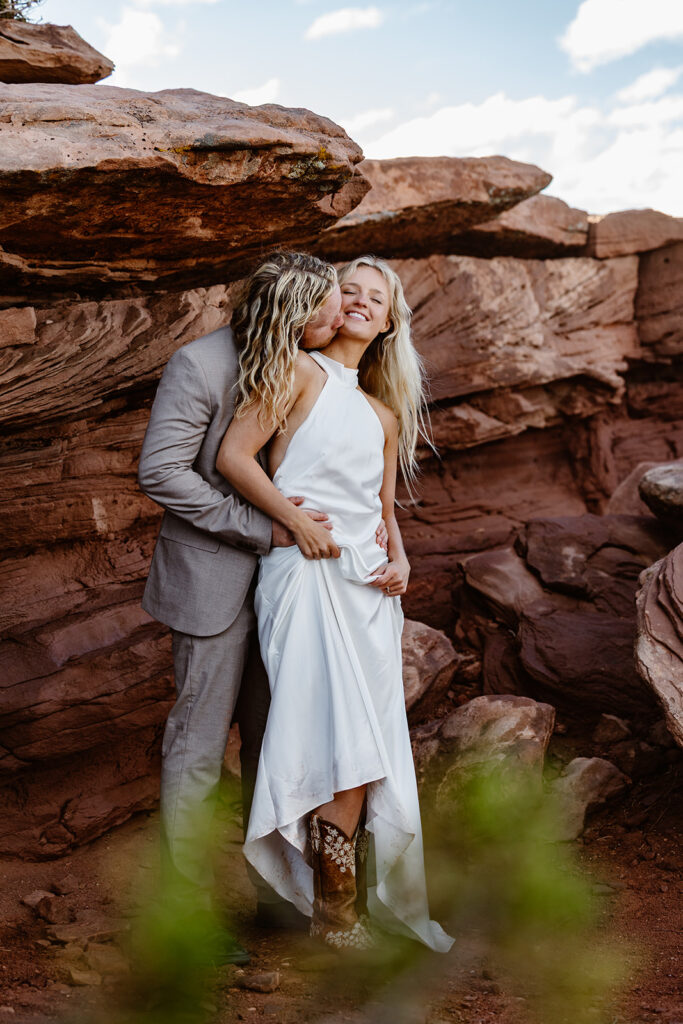 A bride and groom standing in front of some rocks at Dead Horse State Park. The groom is standing behind the bride and snuggling into her. The bride is wearing cowboy boots