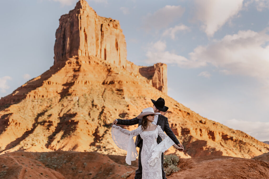 A groom stands behind his bride. Both of their arms are out. They are both wearing cowboy hats, and are standing in front of some rocks at Ida Gulch Trail