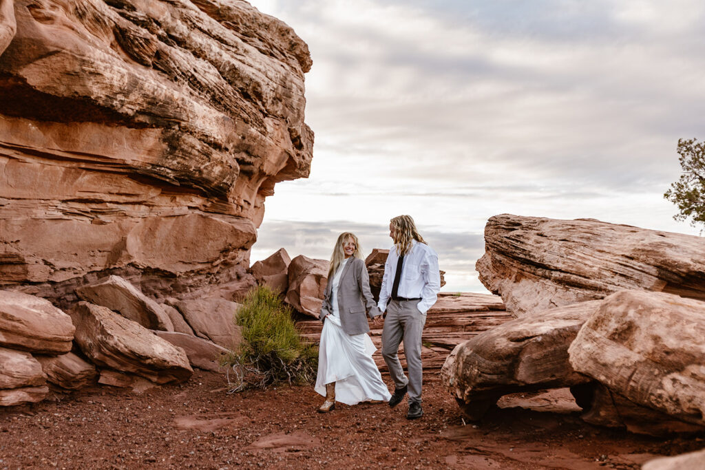 A bride and groom walk through some large red rocks at Dead Horse Point The bride is wearing the grooms jacket because it was chilly at sunrise