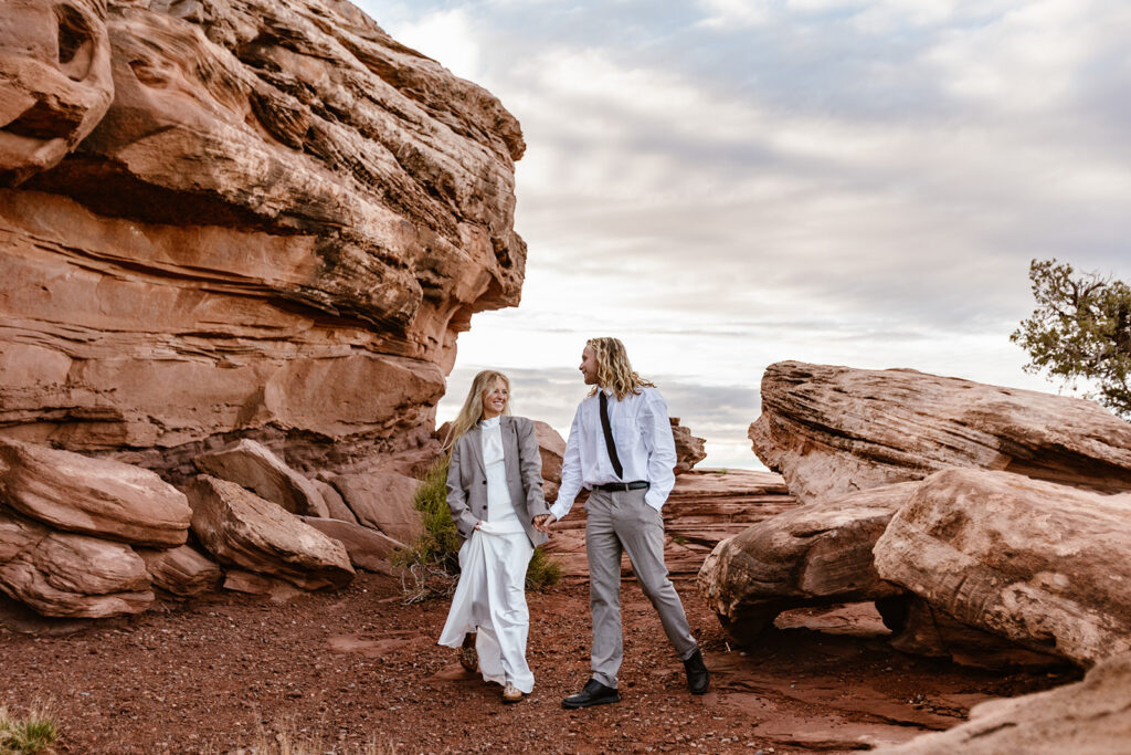 A couple walking at Dead Horse State Park during their elopement. They are holding hands and smiling. The Bride is wearing the grooms coat to help combat the morning chill