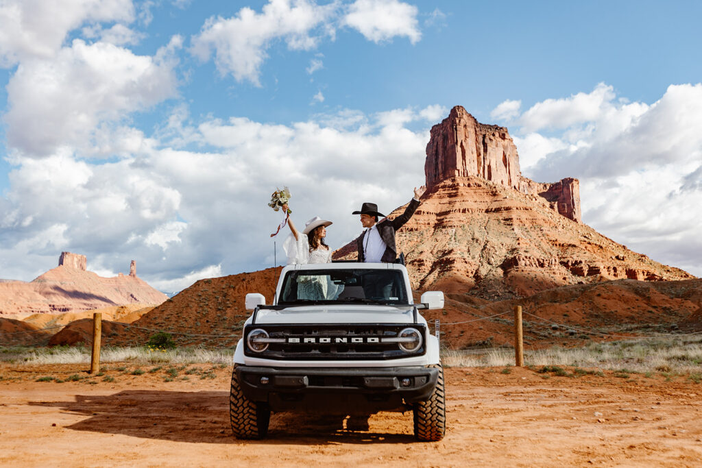 A bride and groom stand in their Bronco's sunroof, with their arms up celebrating their elopement. They are both wearing cowboy hats, and are parked in front of some epic red rocks in Moab