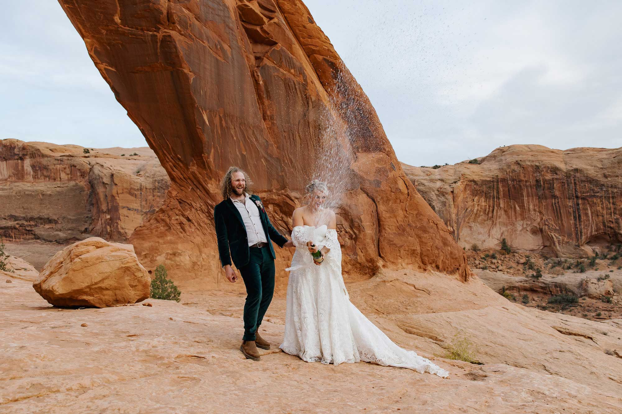 A bride and groom pop champagne in front of the Corona Arch during their adventure elopement in Moab