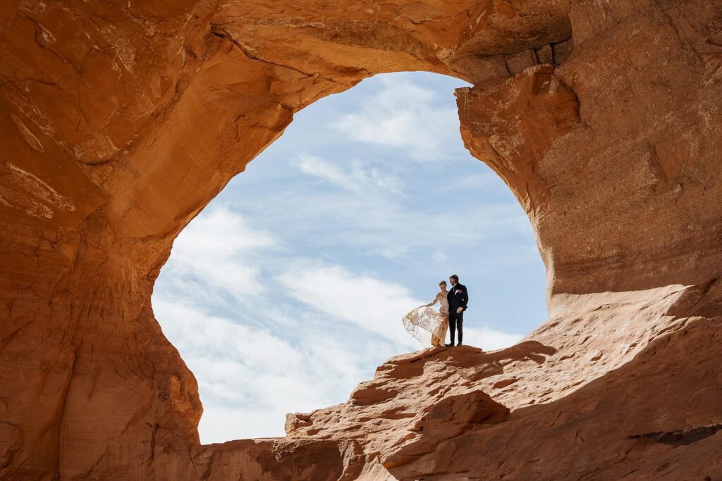 A bride and groom stand in the center of Looking Glass Arch with the blue sky behind them during their elopement in Moab