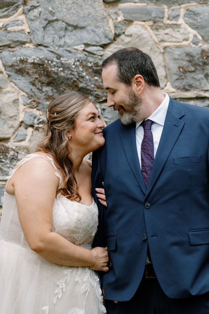 A bride and groom looking at each other smiling. She has her arms wrapped on his, and is leaning into his shoulder. 
