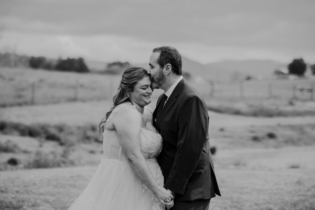 A black and white photos of a groom kissing the forehead of his bride on their elopement day with family.