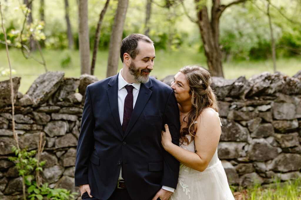 A bride and groom snuggle while looking at each other in front of a stone wall at their AirBnb in Luray  Virginia during their micro wedding