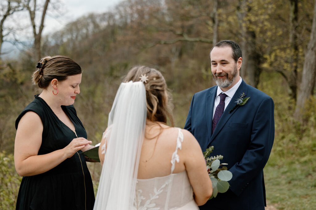 The groom is smiling while the bride reads her personal vows during their wedding ceremony,