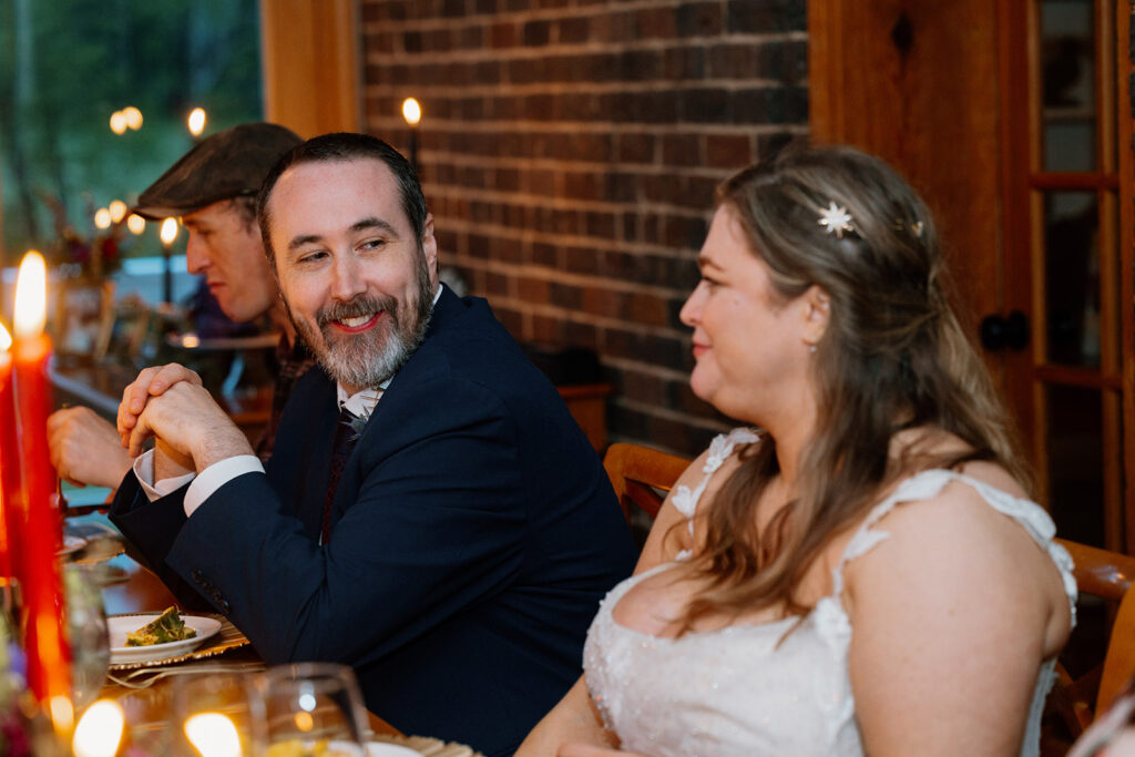 A bride and groom smiling at one another while they sit at the table with their guests at their intimate micro wedding.