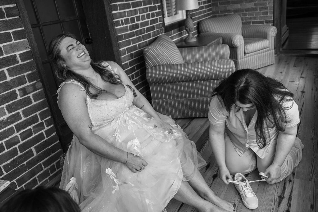 A black and white photo of a bride sitting while her sister puts on her sneakers. The bride is laughing hysterically. 